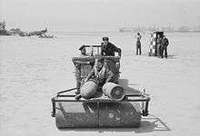  Two men drive a jeep, pulling a ground roller, on which a third man sits. Other men watch in the background. Parked aircraft can be seen in the distance.