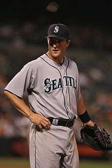 A man in a gray baseball jersey and black baseball cap stands on a grass field wearing a black baseball mitt on his left hand