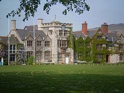 A stone building, partly ivy-covered, seen from a slight angle, with a playing field in the foreground.  The front of the building contains a square battlemented tower, two shaped gables and a series of plain gables.