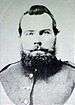 Head of a heavy-set white man with a beard and pointed mustache, wearing a military jacket.