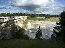 Dam wall shown with water flowing out three of the five floodgates. Spectators are gathered on the viewing area on the top of the wall watching the water flow past.