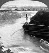 A woman is crossing on a rope over a river.  She wears a wide-brim hat and holds a pole to balance herself while her feet are in buckets.  A double-deck bridge, filled with audience on the lower deck, is in the background.