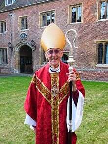 A smiling, bespectacled old man in a red chasuble and gold mitre, holding a crozier and standing on the lawn in front of a large red-brick building.