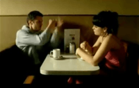A small, square table with round edges against a tan wall. On the table there are coffee cups and a menu, with chairs on two sides, opposite each other. On the right sits a young, brunette woman with large earrings, fringe hair and a pink dress, who stirs the coffee. On the left sits a young man in a grey jumper, who gesticulates the act of punching with his hands.