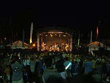 Image of a crowd of people standing behind a music stage at night time watching a woman on bass guitar on the left of the stage (as we view it), a man on drums in the middle of the stage, and another man on guitar on the right of the stage.