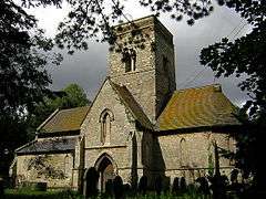 A stone church seen from the south, with a central tower in front of which is a two-storey gabled porch. To the left is a short nave, and to the right an apsidal chancel