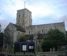 Side view of a substantial stone church with a square clock tower topped with a weather-vane. The tower has two- and three-light arched openings in each face. A triangular-roofed projection to the left has several round-arched windows and a small brown door. The main body of the church, partly obscured by a tree, extends to the right, and has large buttresses topped with spirelets.