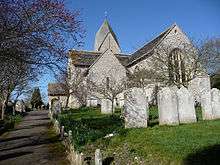 Side view of a flint church surrounded by a graveyard with bare trees and with a path to the left. The top of a tower with a four-sided slate cap is visible behind the main body of the church. A low porch protrudes to the left.