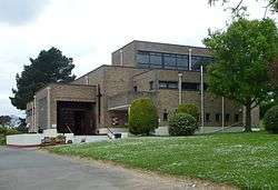 Three-quarter view of a wide, blocky, pale brick building set behind a large grassy area.  There are four sections of different sizes and heights, each with a flat roof.  A wide entrance is in the leftmost projection; on the side wall next to it is a tall crucifix.  White drainpipes are prominent on the exterior.