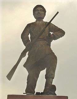 A cement statue depicting a man with a moustache holding a rifle in one hand and a stick in another, wearing traditional clothes. He stands on a polished stone pedestal.