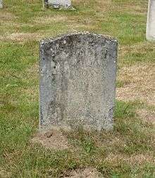 A granite headstone, discoloured by moss, in a grassy churchyard