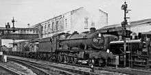 In a black and white photograph, a locomotive (seen from the front and right) is pulling a freight train, of which can be seen a number of open mineral wagons, a closed wagon, and some tankers. The driver is standing at the edge of the cab and seems to be looking at the photographer. The number 4936 is shown on the smoke box at the front, and also the number 681 is written in chalk. Behind the train there is a two storey brick building which includes a covered bridge over the railway. To the right of the locomotive is a second, smaller, tank engine.