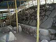 Excavated area with a flat dusty area to the left with some rubble and with steeply sloping well-dressed stonework to the right sat on top of a low vertical stonework base. A ramshackle roof protecting the excavations is supported by wooden poles.