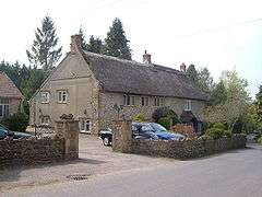 Stone building with thatched roof. In the foreground are cars separated from the road by a stone wall.