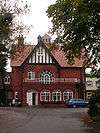 A red brick house, at the end of a long drive, seen from the south. Prominent is a gable with black and white striped pebbledash.