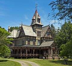 An ornate Victorian house with a peaked central tower seen from a three-quarter angle. It is beige with red trim, with a peaked central tower, polychromatic slate roof and front porte-cochere. There is a curving gravel drive in front and trees on the side; the sky is blue with some clouds.