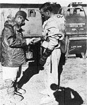 A worker stands and waits while a colleague scans him for signs of radioactivity. Steel tanks with "Crested Ice" painted on them are visible in the background