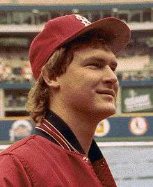 A smiling young man with feathered blond hair wearing a red baseball jacket and red baseball cap