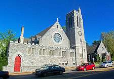 Two pointed gray stone buildings seen from across a street, with cars passing in front and a traffic light visible from the center to the upper left. The one on the left has a tall square stone tower with a smaller tower topped by a cross at the top. The one on the left has a large round window in the middle and a cross at the point on the front. Both have open red doors.