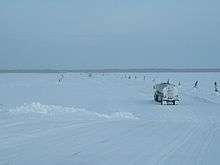 Large trucks, widely spaced, travel single-file across a frozen river.