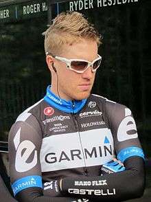 A man in his late twenties wearing a black and white cycling jersey with blue trim, and sun shades.