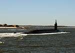 Three quarters view of submarine running on surface near land.