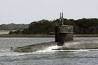 View of front of submarine running on surface, with trees and land in the mid-ground.