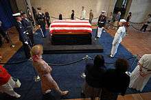 A casket draped with the stripes of the American flag and guarded by military personnel representing each of the five services, as the public files by in single file.