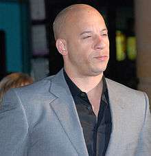 A bald, multi-racial man in a gray suit. Turned at a 45 degree angle from the camera, he squints in the direction of an off-camera light source, which illuminates his face.