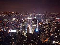 A aerial view of skyscrapers at night. Most of the buildings are lit up with lights coming from the inside of them. In the background, a dark sky with no clouds can be seen.