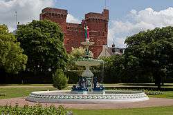 Ornamental fountain in circular pool surrounded by grassy areas. In the background it a red brick building.