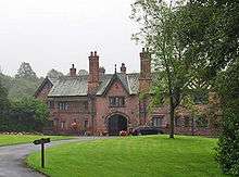 A two-storey house including an archway under a gable and another gable with half-timbering.  In front of it is an area of grass and trees.