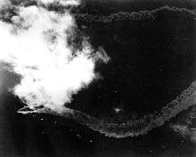 A distant overhead view of a patch of ocean; towards the left, partially obscured by a patch of cloud, is a large warship. The ship's wake trails out in a zig zag pattern behind her.