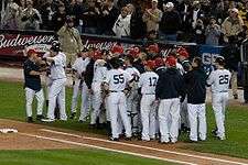 Several men in pinstriped pants and red baseball caps, some wearing pinstriped baseball jerseys and some dark blue-hooded sweatshirts, stand in a group.