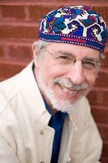 Smiling portrait of Yitzhak Buxbaum c. 2015, wearing glasses, colourful Mizrahi-style kippah, white jacket