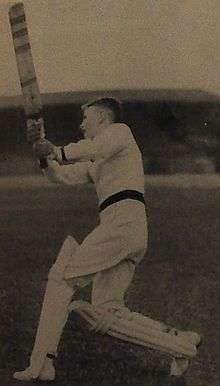 A male youth wearing cricket pads, gloves, white uniform and sweater, stands on the grass and follow through after swinging his bat in a vertical arc.