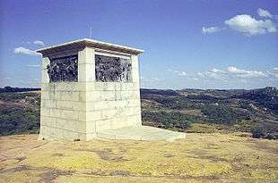 A white, oblong, flat-topped structure, with soldiers depicted on each side in bas relief